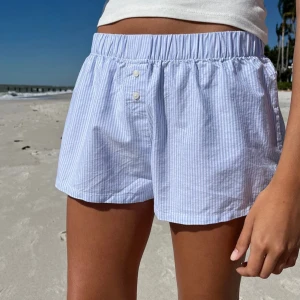 Randiga shorts i bomull - Snygga och bekväma randiga shorts i ljusblått och vitt. De har en elastisk midja och små knappar framtill för en stilren look. Perfekta för en avslappnad dag på stranden eller i parken.