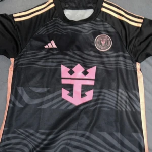 Inter Miami tröja  - Snygg svart fotbollströja från Inter Miami Adidas med korta ärmar och rosa detaljer. Tröjan har ett unikt mönster och klubbmärke på bröstet. Perfekt för fotbollsfans som vill sticka ut på planen.