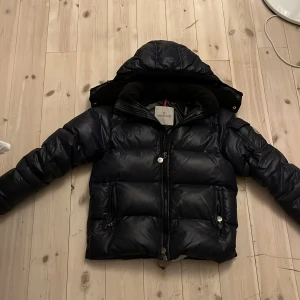 Mörkblå dunjacka från Moncler - Säljer en snygg mörkblå dunjacka från Moncler med huva och dragkedja. Jackan har Moncler-logga på ärmen och en unik serietidningsetikett på insidan. Perfekt för kalla dagar!