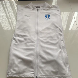 Vit jacka från Puma med Malmö FF-logga - Säljer en stilren vit jacka från Puma med Malmö FF-logga på bröstet. Jackan har dragkedja framtill och är långärmad. Perfekt för fotbollsfans som vill visa sitt stöd. 🩵