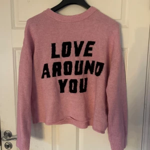 Rosa stickad tröja med text - Säljer en rosa stickad tröja med texten 'LOVE AROUND YOU' i svart på framsidan. Tröjan har långa ärmar och en avslappnad passform. 