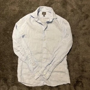 Ljusblå linneskjorta från H&M - Snygg ljusblå skjorta i linne från H&M. Perfekt för varma dagar med sin lätta och luftiga känsla. Skjortan har långa ärmar och knappar framtill. En klassisk och stilren design som funkar till många tillfällen. Endast använt den vid ett tillfälle så skick som nytt.