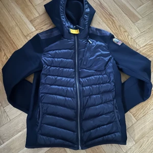 Marinblå cardigan från Parajumpers - Säljer en marinblå cardigan från Parajumpers med huva och dragkedja. Jackan har quiltade paneler fram och bak, samt en logga på ärmen.