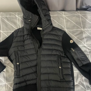 Svart dunjacka från Moncler - Snygg svart dunjacka från Moncler med huva och dragkedja. Jackan har ribbade detaljer och är perfekt för kyligare väder. Den har även praktiska fickor med knappar och en logga på ärmen.