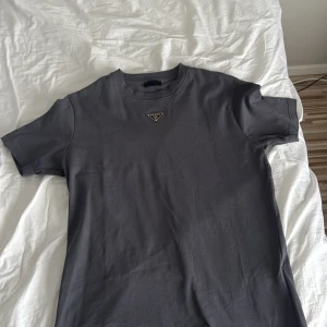 Grå t-shirt från Prada - Snygg grå t-shirt från Prada med en diskret logga på bröstet. Perfekt för en stilren look. T-shirten har en klassisk passform och är kortärmad.
