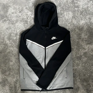 Nike tech fleece  - Säljer en snygg huvtröja från Nike i svart och grått med dragkedja och huva. Tröjan har ett stilrent mönster med vita detaljer och Nike-logga på bröstet. Perfekt för en sportig look eller avslappnad stil.