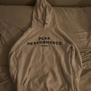 Vit hoodie från Peak Performance - Säljer en stilren vit hoodie från Peak Performance med svart text på bröstet. Den har en klassisk design med en stor ficka framtill och justerbar huva med snören. Perfekt för en avslappnad look.