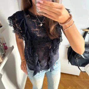 Svart spetsblus - Elegant svart spetsblus med korta ärmar och vackra spetsdetaljer. Perfekt för en stilfull look. Blusen har en rund hals och är lätt transparent, vilket ger en sofistikerad touch.