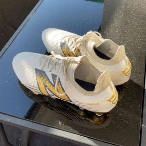 Endrick new balence limited edition fotbollsskor - Snygga vita fotbollsskor med guld och svart detaljer. Skorna har snörning och en modern design med en stor logotyp på sidan. Perfekta för fotbollsplanen med en stilren look.