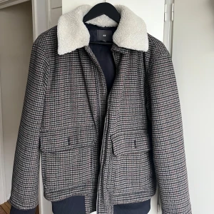 Rutig jacka med teddykrage från H&M - Snygg rutig jacka från H&M med en mysig vit teddykrage. Jackan har långa ärmar och stängs med dragkedja. Perfekt för kyliga dagar med sin stilrena design och praktiska fickor framtill.