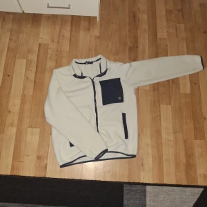 Vit fleecejacka med blå detaljer - Säljer en vit fleecejacka med blå detaljer och dragkedja. Jackan har en bröstficka och två sidofickor, alla med blå kant. Perfekt för kyliga dagar!