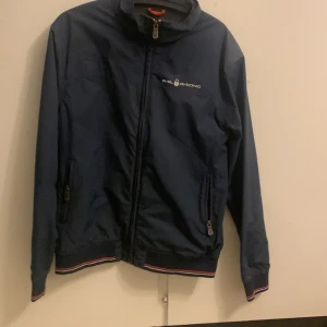 Mörkblå jacka från Sail Racing - Säljer en mörkblå jacka från Sail Racing med dragkedja och ribbade detaljer vid ärmslut och nederkant. Jackan har långa ärmar och en stilren design med logotyp på bröstet och ryggen. Perfekt för en sportig look!