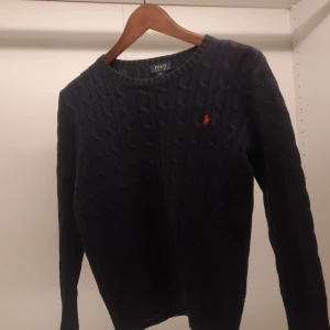 Mörkblå stickad tröja från Ralph Lauren - Säljer en snygg mörkblå stickad tröja från Ralph Lauren med kabelstickat mönster. Tröjan har en liten röd logotyp på bröstet och långa ärmar. Perfekt för en stilren look!