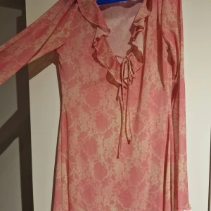 Rosa mönstrad mesh klänning med volangdetaljer - rosa mesh klänning med ett subtilt mönster och volangdetaljer vid halsen perfekt för sommarn och stranden. Klänningen har långa ärmar och en knytning framtill som ger en romantisk touch. Perfekt för en somrig look.