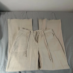 Beige linnebyxor med snörning - Snygga beige linnebyxor med avslappnad passform och snörning i midjan. Byxorna har både fram- och bakfickor samt en knappstängning. Perfekta för en ledig stil.