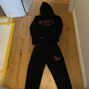 Svarta mjukisbyxor från Supply & Demand - Svarta mjukisbyxor med snörning i midjan och ett coolt tryck på benet. Byxorna har ett mönstrat band längs sidorna som ger en extra touch. Perfekta för en avslappnad stil.