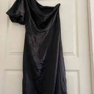 Svart asymmetrisk klänning från Boohoo - Snygg svart asymmetrisk klänning från Boohoo i satin med puffärm. Perfekt för en elegant look med sin unika design och glansiga material. Klänningen har en oneshoulder-stil som ger en modern touch.