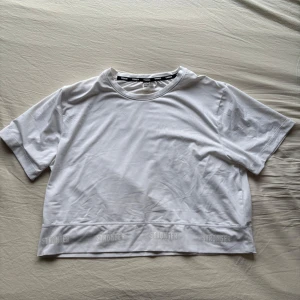 Vit t-shirt från Stronger - Säljer en vit t-shirt från Stronger i storlek medium. T-shirten är tillverkad av 90% polyester och 10% elastan, vilket ger en skön och stretchig passform. Den har korta ärmar och ett band med logotypen 'STRONGER' längs fållen.