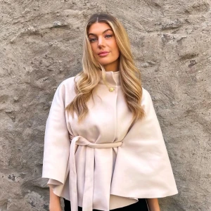Beige kappa med knytband - Snygg beige kappa med knytband i midjan och vida ärmar. Perfekt för en stilren look. Den har en elegant design med en knappdetalj vid halsen.