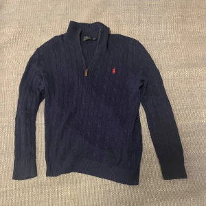 Mörkblå kabelstickad halvzip från Polo Ralph Lauren - Säljer en stilren mörkblå kabelstickad tröja från Polo Ralph Lauren. Tröjan har en dragkedja vid halsen och det ikoniska röda logotypbroderiet på bröstet. Perfekt för en klassisk och avslappnad look. Kablarna är lite slitna så om man har en ljus tröja under kan det synas igenom lite. Pris kan diskuteras vid snabb affär!