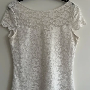 Vit spetsklänning med v-ringning - Charmig vit spetsklänning med v-ringning och korta ärmar. Perfekt för somriga dagar eller speciella tillfällen. Den har ett vackert blommigt mönster i spetsen som ger en romantisk känsla.