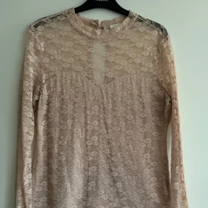 Beige/rosa spetsblus - Elegant beige och rosa spetsblus med långärmad design och en vacker öppen ryggdetalj. Perfekt för att ge en romantisk touch till din outfit. Blusen har en hög hals och ett delikat blommönster.