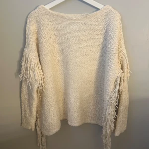 Beige stickad tröja från Ralph Lauren - Säljer en snygg beige stickad tröja från Ralph Lauren med fransdetaljer längs ärmarna. Tröjan har en avslappnad passform och långa ärmar, perfekt för kyligare dagar. Ett stilrent plagg som ger en bohemisk touch till din garderob.