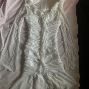 Student klänning med volangdetaljer - Säljer en elegant vit klänning med volangdetaljer och långa ärmar. Klänningen har en v-ringad design med knytning framtill och en figurnära passform. Perfekt för speciella tillfällen!                                                                      Har bara använd den en gång vid min studentmottagning.