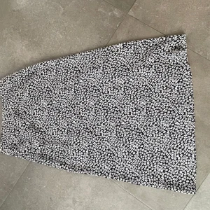 Svartvit blommig kjol med slits - Snygg svartvit kjol med blommigt mönster och en stilren slits på sidan. Perfekt för en chic look. Kjolen har en omlott-design som ger en fin passform.