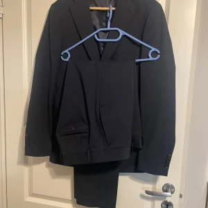 Svart kostym från Pour Homme - Säljer min Pour Homme kostym från Strömsons då jag har växt ur den.   Den är endast använd ett mycket fåtal gånger - på studenten och någon enstaka gång efteråt.   För storleksreferens var jag strax under 180cm lång och vägde mellan 70-75 när kostymen användes. Storleken på kavajen och byxorna är B48.  Det går jättebra att prova kostymen innan köp!