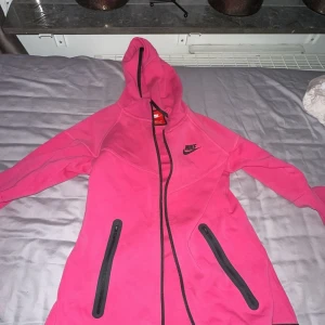 Rosa hoodie från Nike - Säljer en snygg rosa hoodie från Nike med dragkedja och huva. Jackan har svarta detaljer och två praktiska fickor med dragkedjor framtill. Perfekt för en sportig look eller en avslappnad dag.