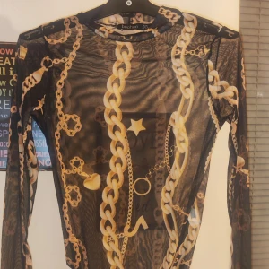 Svart mesh body med kedjemönster från Boohoo - Säljer en svart mesh body från Boohoo med ett coolt kedjemönster i guld. Toppen har långärmade ärmar och en tight passform, perfekt för en trendig look. Den är genomskinlig och ger en edgy stil.