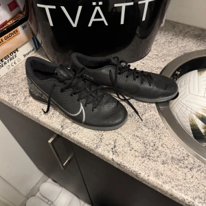 Svarta Nike fotbollsskor inomhus/futsal - Snygga svarta Nike fotbollsskor med vit swoosh-logga. Skorna har snörning och är perfekta för fotbollsplanen. De har en stilren design och är gjorda för att ge bra grepp och komfort. 