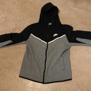 Grå och svart hoodie från Nike storlek 158cm - 170cm - Snygg grå och svart hoodie från Nike med dragkedja och huva. Perfekt för en sportig look. Den har långa ärmar och en vit Nike-logga på bröstet.