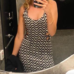 Svartvit klänning  - Trendig svartvit klänning med zigzag-mönster och asymmetrisk fåll. Klänningen har en djup urringning och sidodetaljer med utskärningar. Perfekt för en stilfull look.