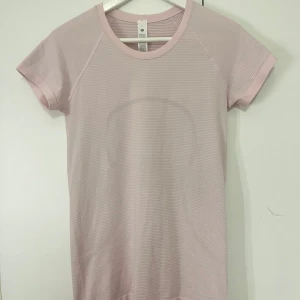Ljusrosa kortärmad topp från Lululemon - Säljer en ljusrosa kortärmad topp från Lululemon med diskret randigt mönster. Perfekt för träning eller en avslappnad dag. Toppen har en rund halsringning och är tillverkad i ett mjukt och stretchigt material. 