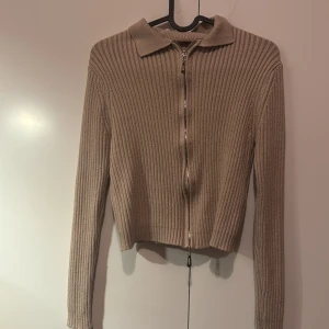 Beige ribbad kofta med dragkedja - Säljer en stilren beige ribbad kofta med dragkedja och krage. Perfekt för lager-på-lager under kyligare dagar. Långärmad och croppad design som ger en modern look.