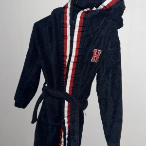 Mörkblå morgonrock från Tommy Hilfiger - Säljer en mörkblå morgonrock från Tommy Hilfiger med röd och vit randig detalj längs med framsidan. Den har en broderad bokstav 'H' på bröstet och ett bälte i midjan. Perfekt för att mysa hemma! Storleken är Junior L.