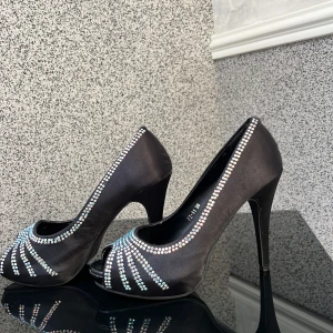 Svarta pumps med strassdetaljer - Eleganta svarta pumps med hög klack och glittrande strassdetaljer. Perfekta för en festlig kväll eller speciella tillfällen. Skorna har en öppen tå och är tillverkade i ett satinliknande material.