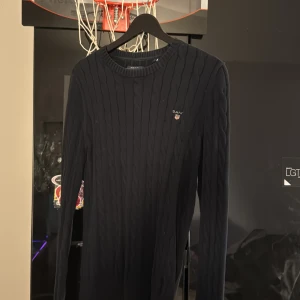 Mörkblå stickad tröja från GANT - Säljer en stilren mörkblå stickad tröja från GANT i premium bomull. Tröjan har ett diskret kabelstickat mönster och rund halsringning. Perfekt för kyligare dagar.