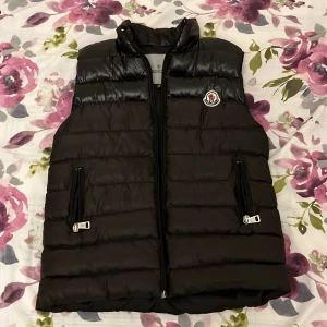 Svart dunväst från Moncler - Säljer en snygg svart dunväst från Moncler med dragkedja och broderad logga på bröstet. Västen har en quiltad design och två fickor med dragkedja. Perfekt för kyligare dagar.