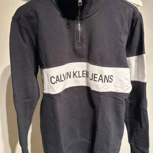 Svart tröja från Calvin Klein Jeans - Snygg svart tröja från Calvin Klein Jeans med vit panel och logga över bröstet. Tröjan har en halv dragkedja och långa ärmar, perfekt för en stilren look.nypris 1500kr