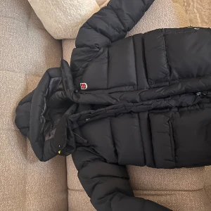 Mörkblå dunjacka från Fjällräven - Säljer en svart dunjacka från Fjällräven med huva och dragkedja. Jackan har en pufferdesign och är perfekt för kalla dagar. Den har justerbara detaljer och en liten logotyp på bröstet.