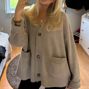 Beige kofta med knappar - Mysig beige kofta med stora knappar och två fickor framtill. Den har en loose passform och långa ärmar, perfekt för kyliga dagar. Ribbstickade detaljer vid ärmslut och nederkant.