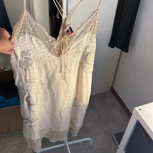 Beige klänning från Zadig & Voltaire - Elegant beige klänning med spetsdetaljer och subtila glittriga inslag. Klänningen har tunna axelband och ett vackert mönster med broderade detaljer. Perfekt för en somrig look.
