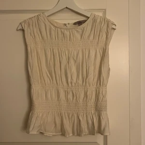 Vit ärmlös blus från H&M - Söt krämvit blus från H&M med smockdetaljer och volang nedtill, använd fåtal gånger så i väldigt bra skick 