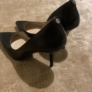 Svarta klackar från Michael Kors - Eleganta svarta klackar från Michael Kors med spetsig tå och klack. Skorna har en stilren design med en dekorativ detalj på hälen. Perfekta för en chic look.