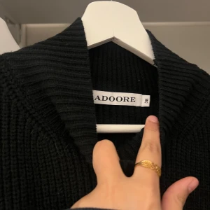 Svart stickad klänning från Adoore - Snygg svart stickad klänning från Adoore i storlek 38. Klänningen har en figurnära passform med långa ärmar och en ribbad struktur som ger en stilren look. Perfekt för kyligare dagar.