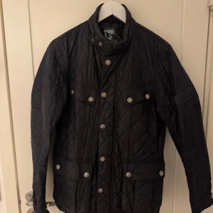 Barbour Svart quiltad jacka med knappar - Snygg svart quiltad jacka med knappar och hög krage. Perfekt med sin stilrena design och praktiska fickor. Passar bra till både vardag och mer uppklädda tillfällen. Nypris: ~3000kr