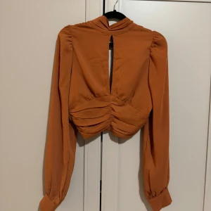 Orange knytblus med långa ärmar - Snygg orange blus med knytning framtill och långa ärmar. Blusen har en öppen rygg och knappar vid ärmsluten. Perfekt för en stilren look.